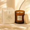 scentulp-geurkaars-ylang-blossom-340-g.webp Amberkleurige geurkaars in glazen pot naast een crèmekleurige doos en houten deksel.